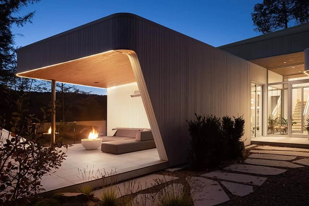 Illuminated covered outdoor patio at night with angled wood soffit, fire bowl, and chaise lounge