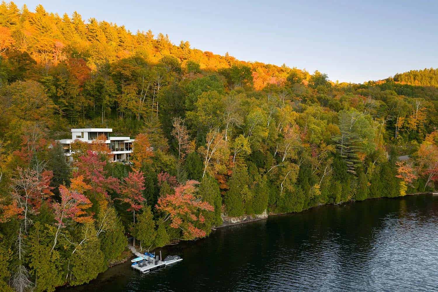 Aerial view of contemporary white home nestled in vibrant autumn forest above a dark lake with private dock