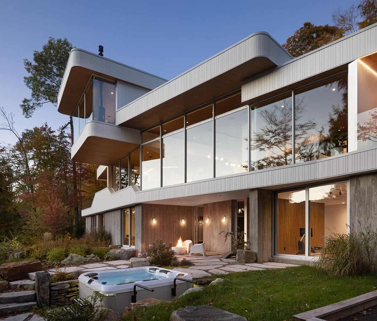 Rear exterior at dusk with hot tub, stone patio, fire feature, and illuminated multi-level glazed facade
