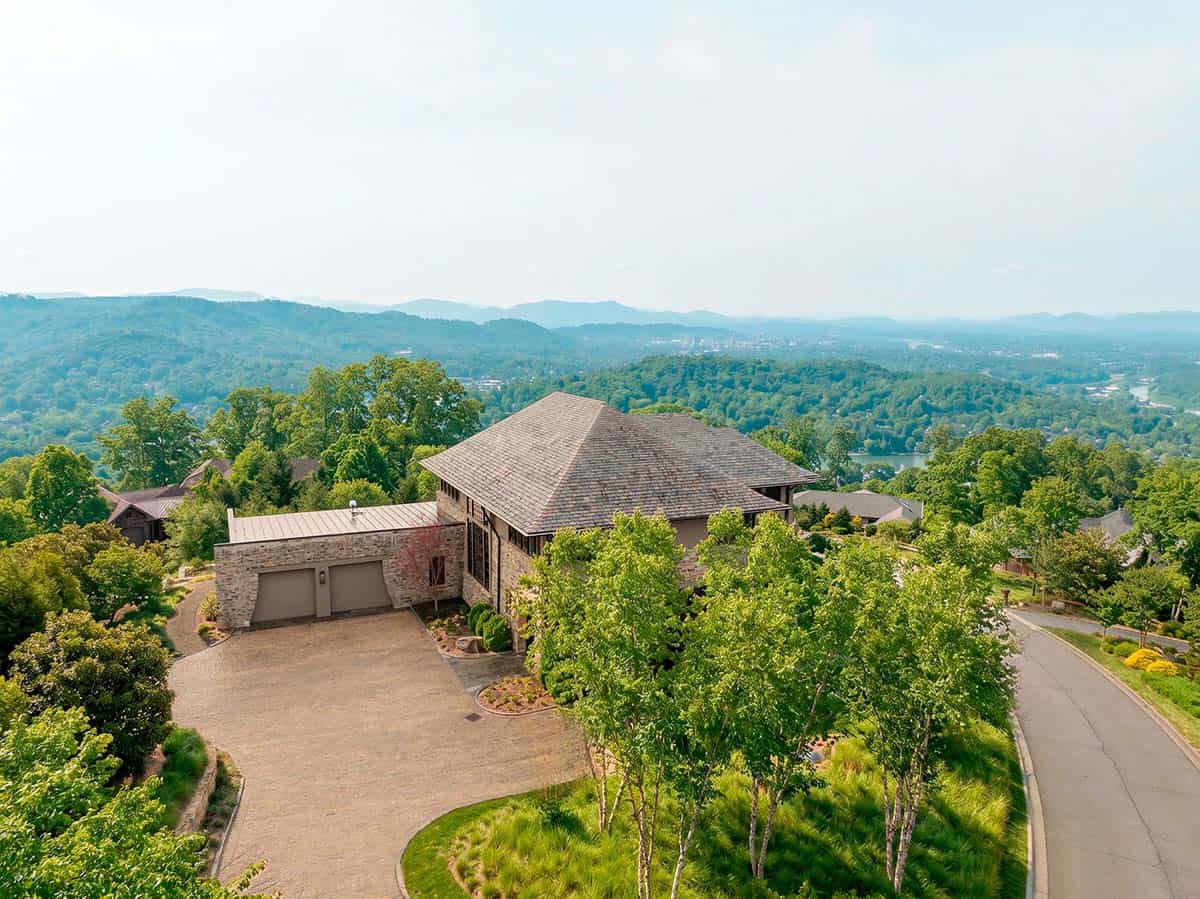 Aerial view of a contemporary stone and stucco home with a hip roof and two-car garage, surrounded by trees with mountain views in the distance, North Carolina