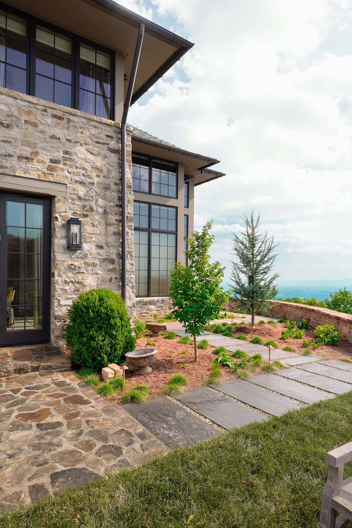 Side exterior of a stone home with large black-framed grid windows, a flagstone pathway, and layered landscaping with young trees and shrubs overlooking a mountain view
