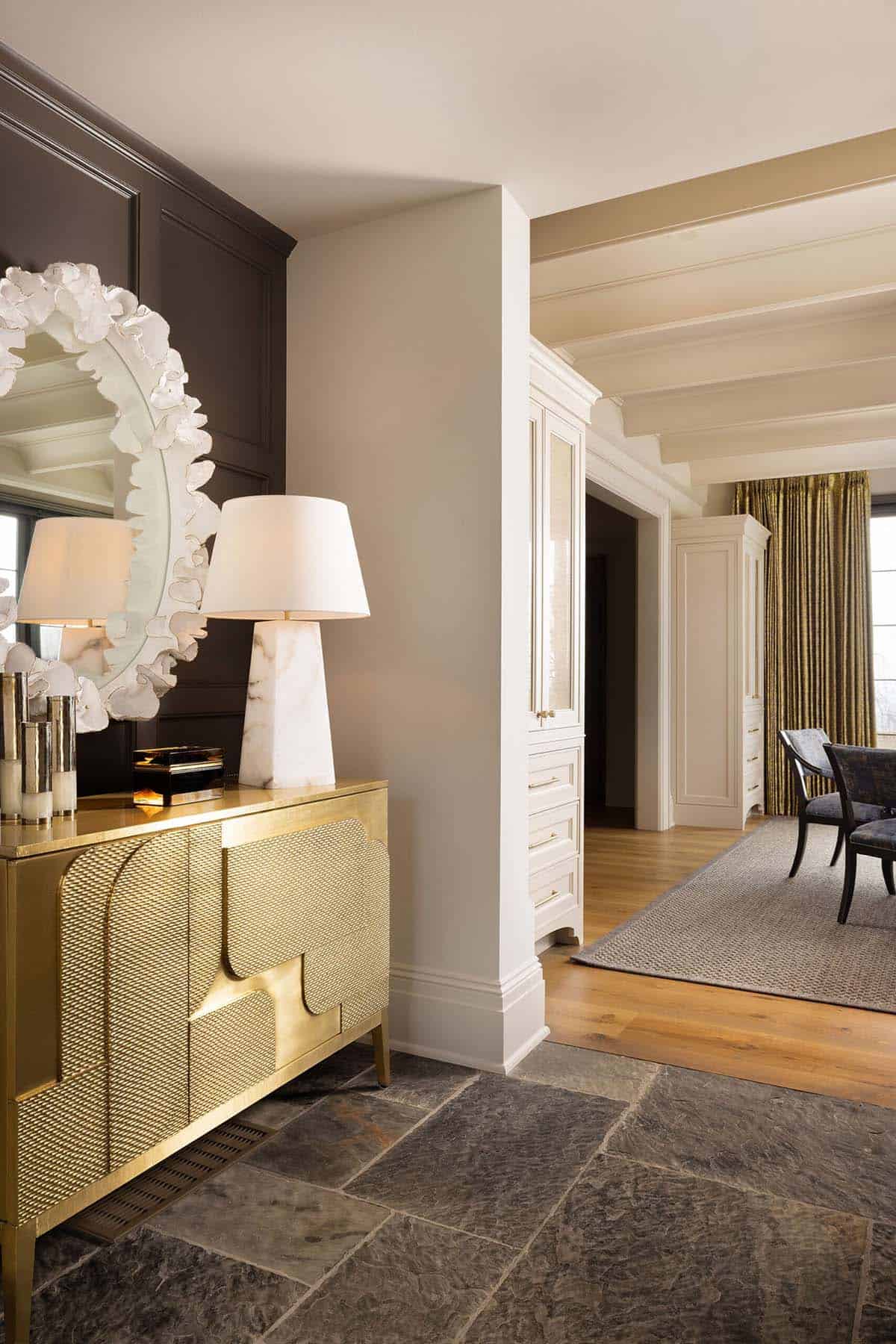 Entry hall with a gold textured sideboard, sculptural white floral mirror, marble table lamp, dark paneled walls with picture frame molding, and a view into an adjoining room with wood floors and gold drapery