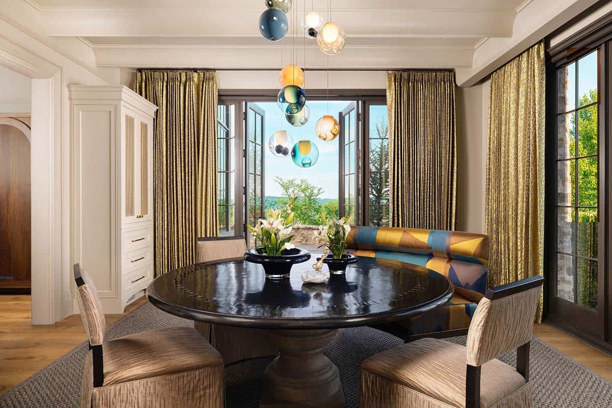 Breakfast nook with a dark round pedestal table, upholstered chairs, a colorful geometric banquette, multi-colored blown glass pendant cluster, and casement windows open to a wooded view