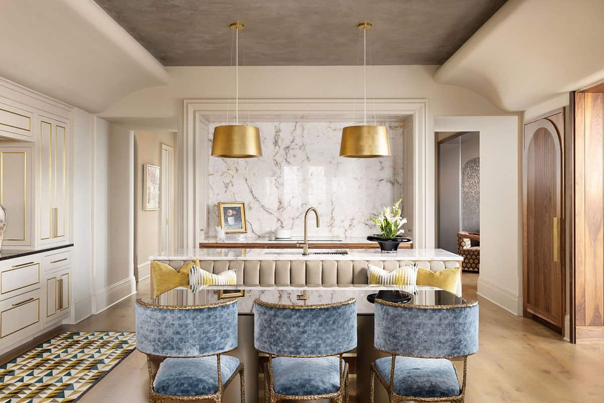 Kitchen with a large marble island topped with a waterfall edge, blue velvet barstools with brass frames, gold drum pendant lights, white cabinetry with gold trim, and a full-height marble backsplash