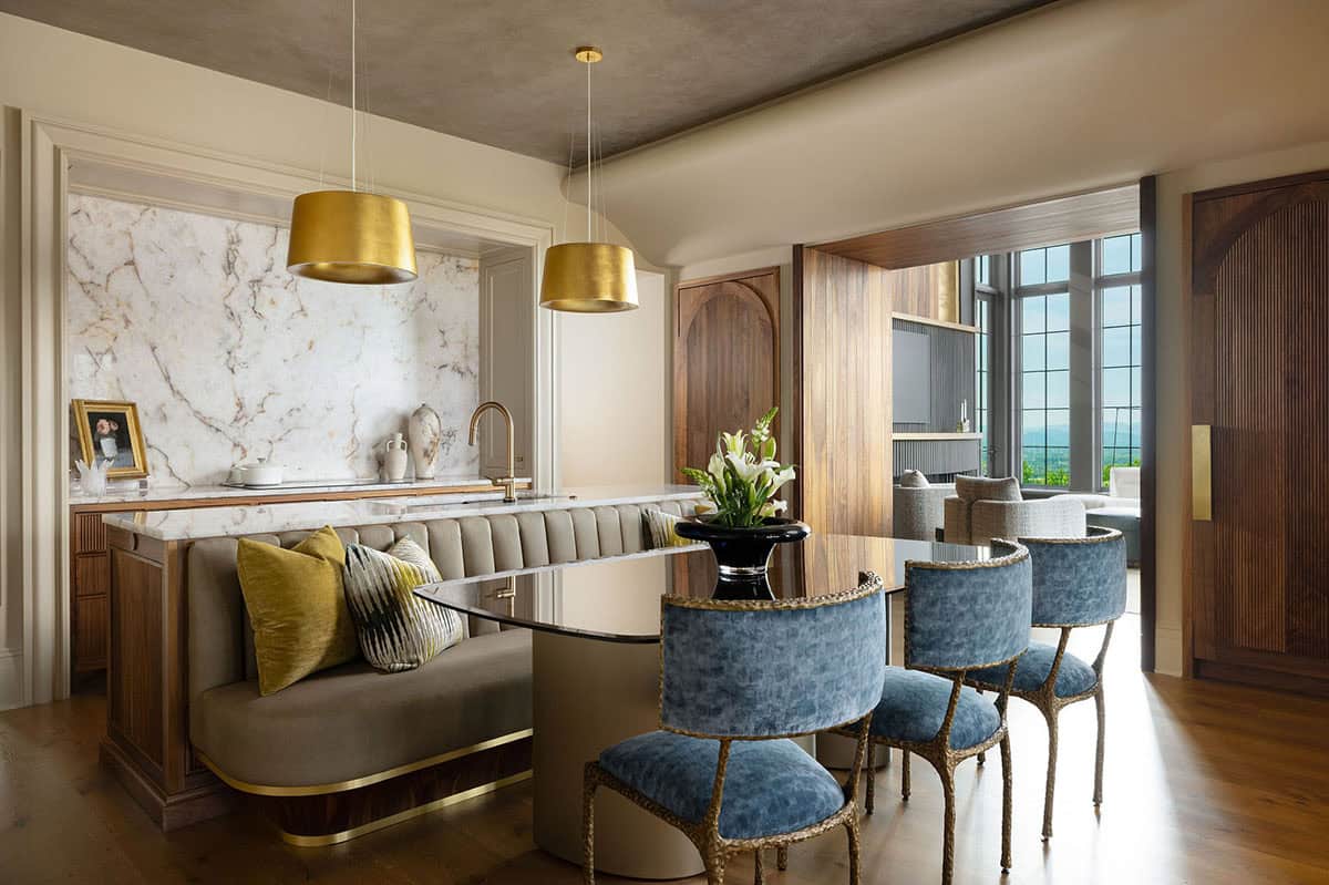 Kitchen island with a built-in upholstered banquette, blue velvet chairs, marble countertop, gold pendant lights, and a view through walnut arched doors into a sitting room with mountain views
