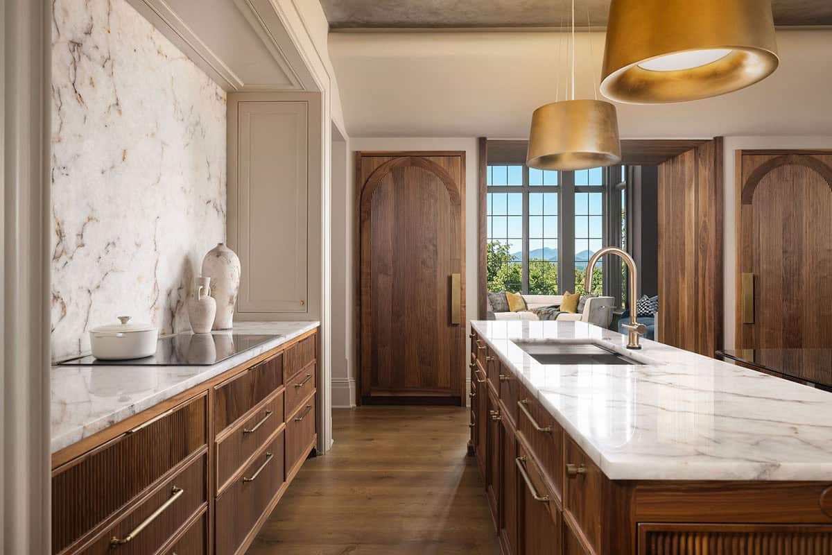 Kitchen with fluted walnut cabinetry, a marble countertop and backsplash, brass hardware, a farmhouse sink in the island, and a walnut arched door leading to a sitting room with grid windows