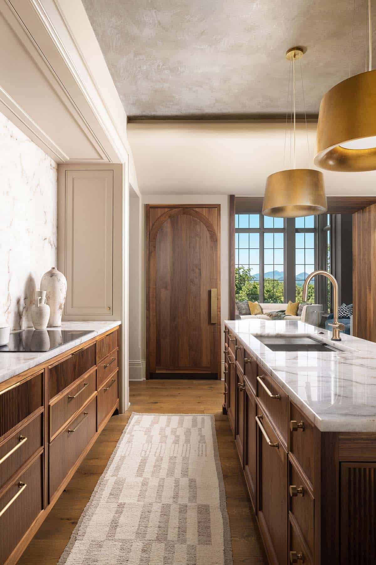 Kitchen galley view showing fluted walnut lower cabinets, a marble island with undermount sink, gold drum pendants, a textured plaster ceiling, and mountain views through large grid windows