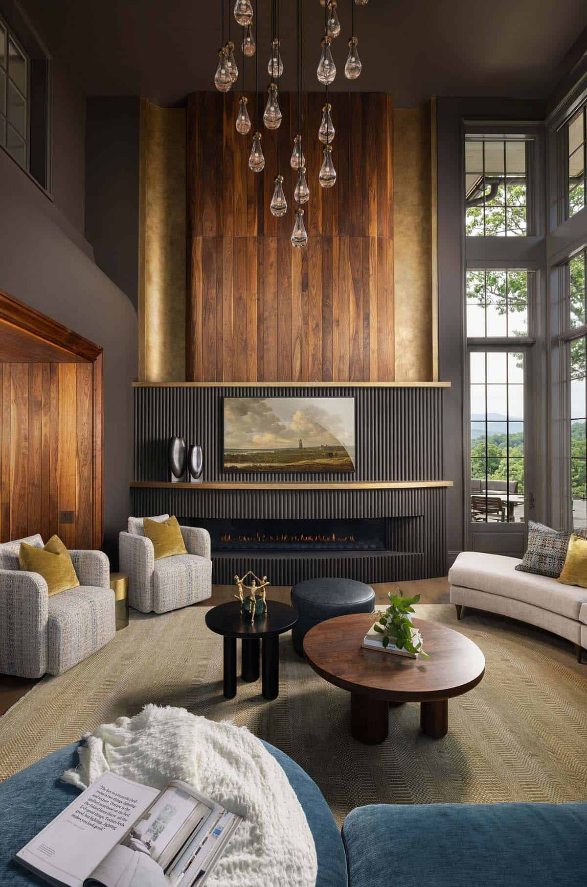 Double-height living room with a dramatic walnut and brass fireplace surround, fluted charcoal firebox, linear gas fireplace, crystal raindrop pendant chandelier, and mixed seating arrangement on a herringbone rug