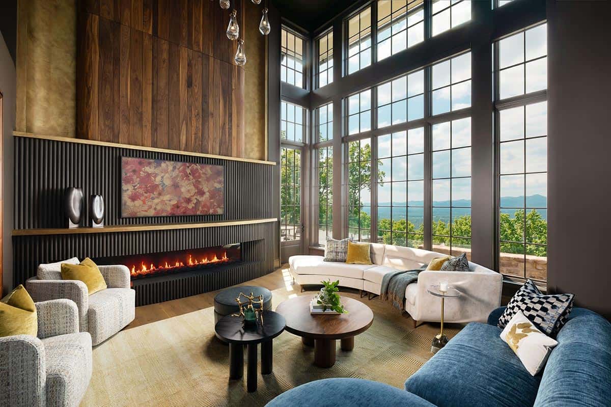 Double-height living room with floor-to-ceiling grid windows framing Blue Ridge Mountain views, a linear gas fireplace with fluted charcoal surround, walnut and brass wall paneling, and curved seating in cream and blue
