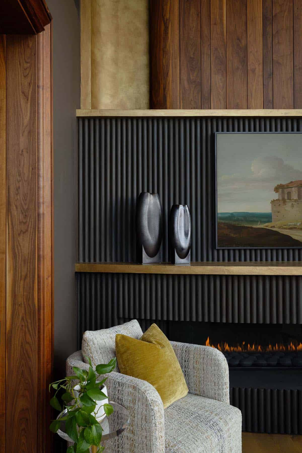 Close-up of a fluted charcoal fireplace surround with brass shelf ledge, two dark sculptural glass vases, a framed landscape painting on a smart display, and walnut paneling above