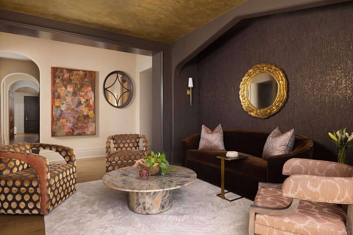 Jewel-toned sitting room with a gold leaf ceiling, dark textured wallcovering, brown velvet curved sofa, gold sunburst mirror, bold patterned armchairs, and a round marble coffee table