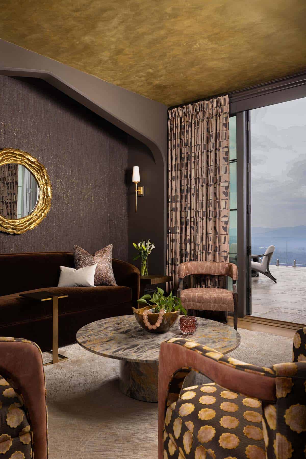 Sitting room with a gold leaf ceiling, dark textured wallcovering, brown velvet sofa with mixed throw pillows, round marble coffee table, and sliding glass doors opening to a terrace with mountain views