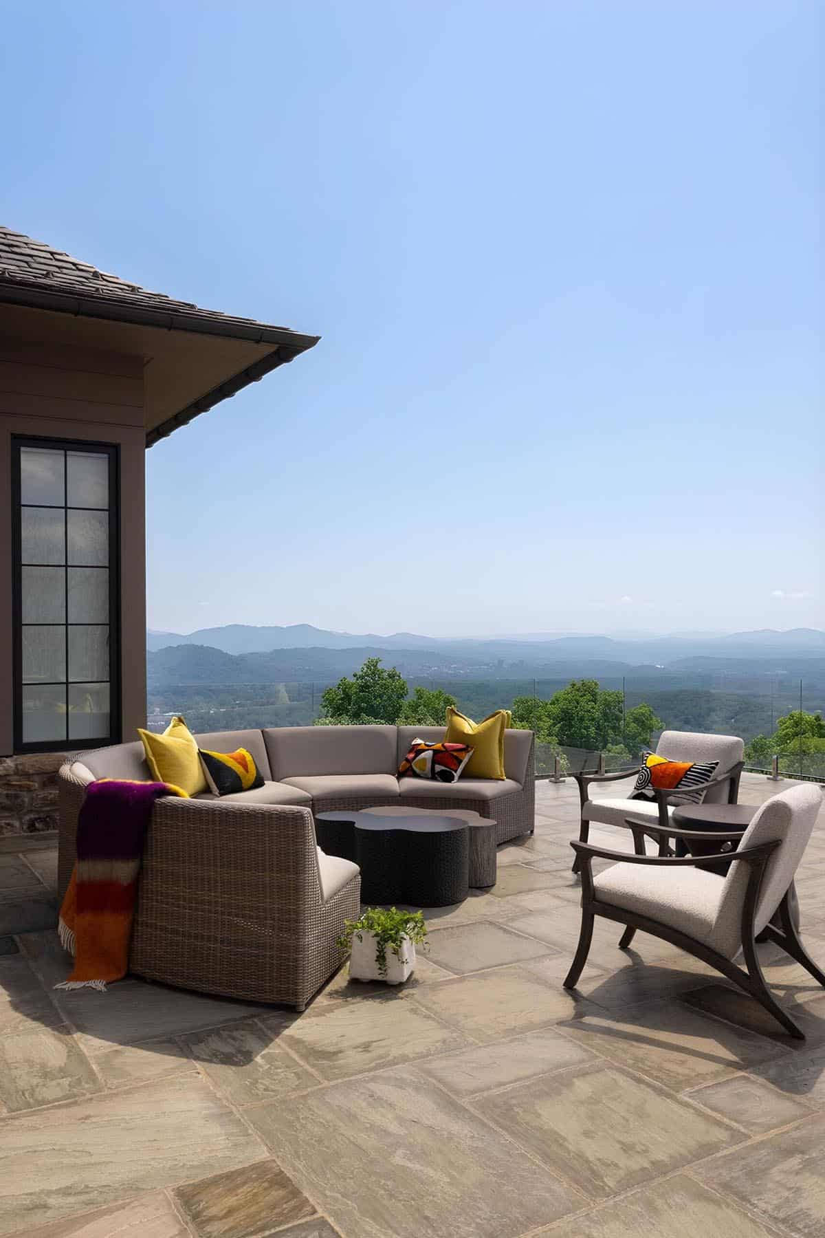 Stone-paved terrace with a curved wicker sectional, colorful accent pillows, dark organic coffee table, and open mountain views under a clear blue sky