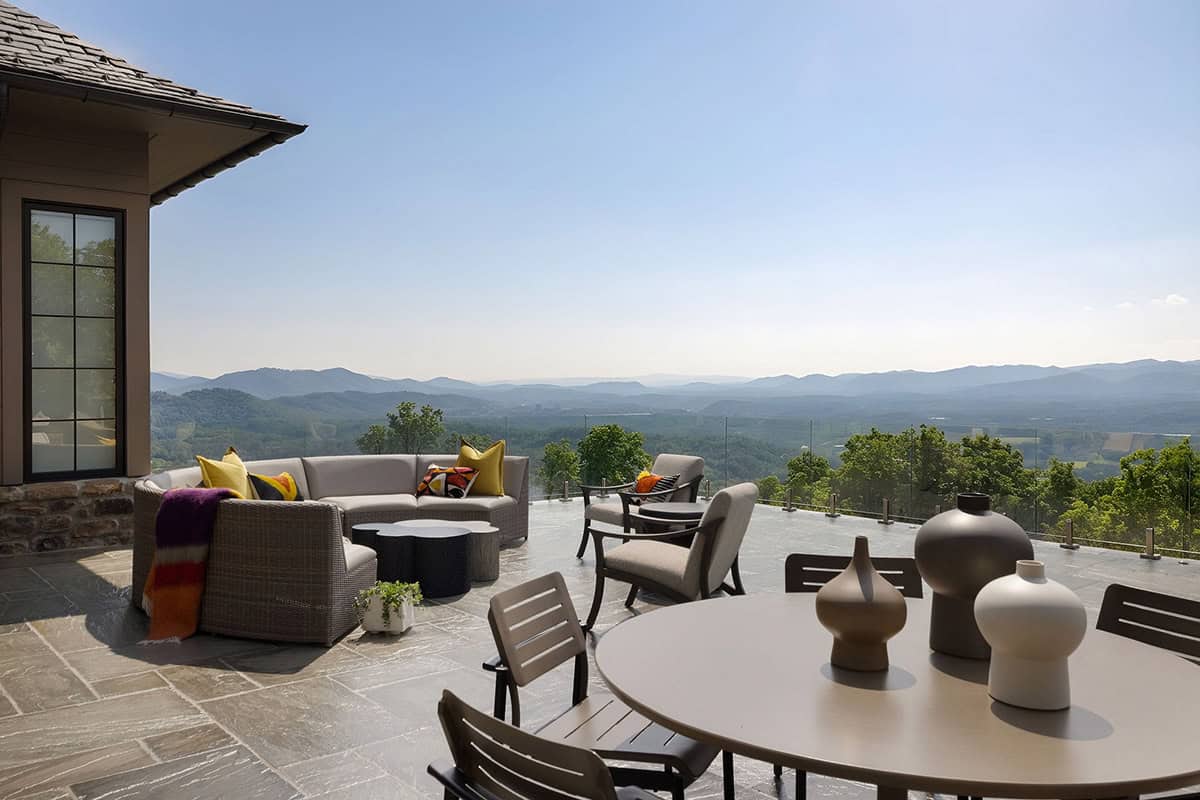 Expansive stone terrace with a wicker sectional, outdoor dining set, decorative ceramic vases, glass panel railing, and sweeping Blue Ridge Mountain views