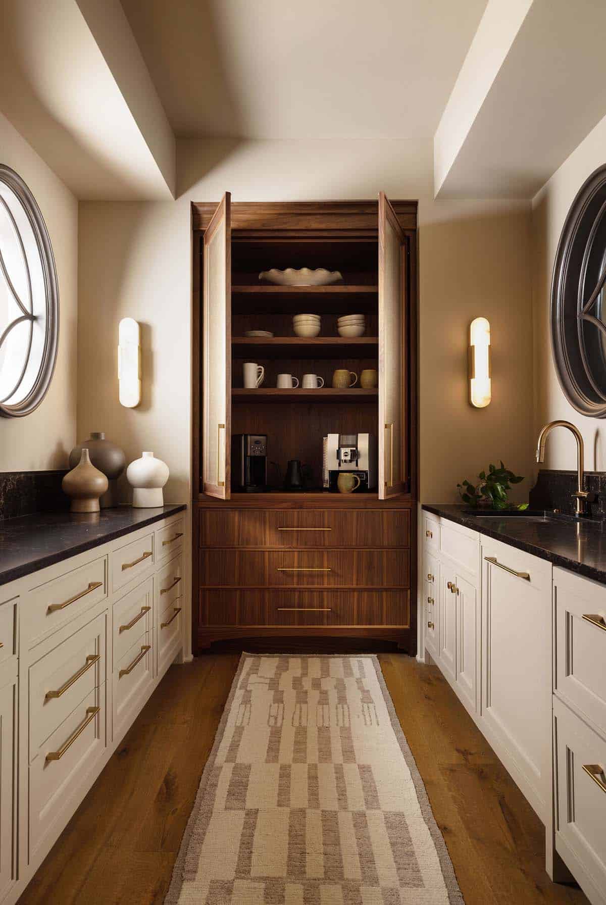 Butler's pantry with white cabinetry, black stone countertops, brass hardware, a tall open walnut cabinet displaying dishware and coffee appliances, flanked by oval windows and wall sconces
