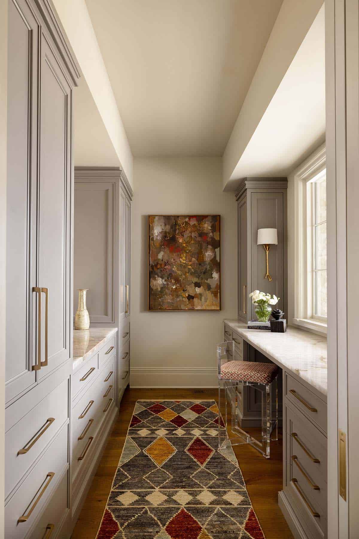 Walk-in closet with floor-to-ceiling gray painted cabinetry, brass hardware, a marble vanity counter with lucite stool, abstract oil painting, and a kilim runner on wood floors