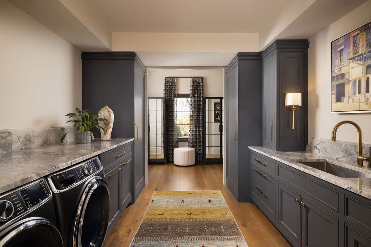 Laundry room with dark gray shaker cabinetry, marble countertops, brass hardware, front-load washer, undermount sink with brass faucet, and a colorful runner on wide-plank oak floors