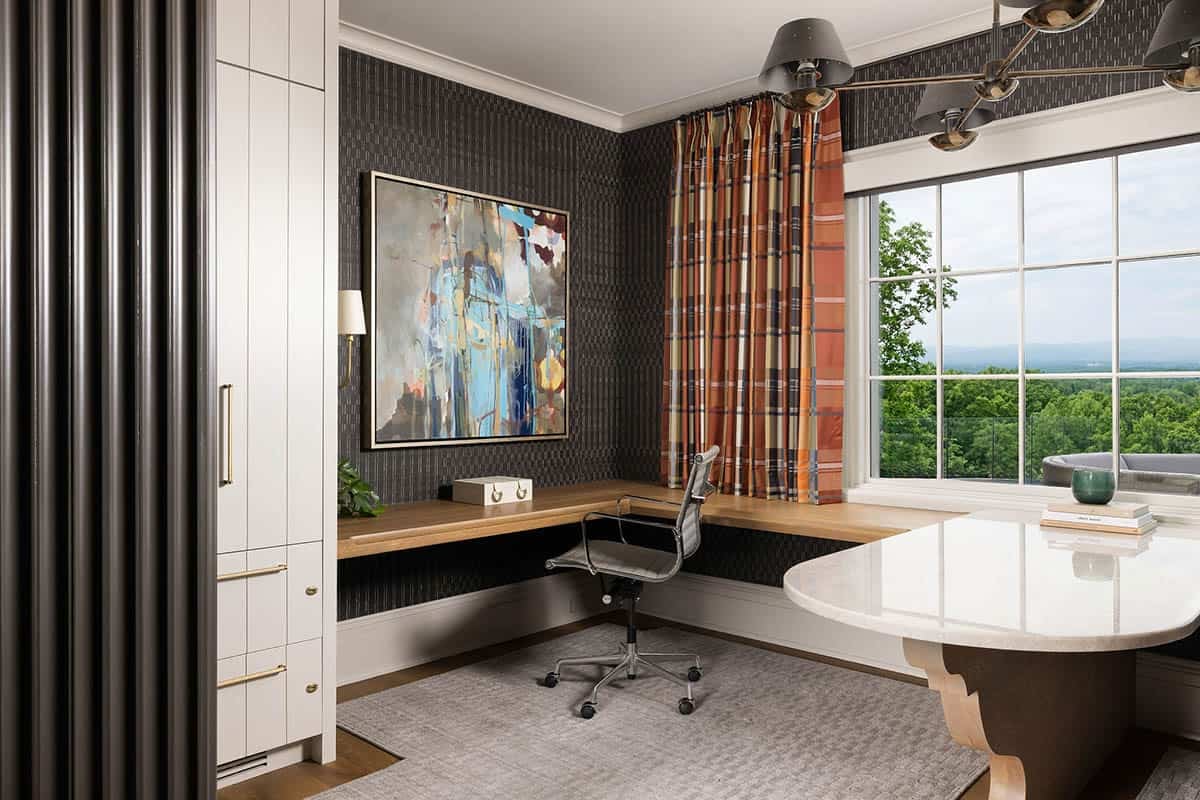 Home office with dark textured wallcovering, a floating wood L-shaped desk, abstract canvas painting, plaid drapery, large grid window with mountain views, and a marble-topped desk in the foreground