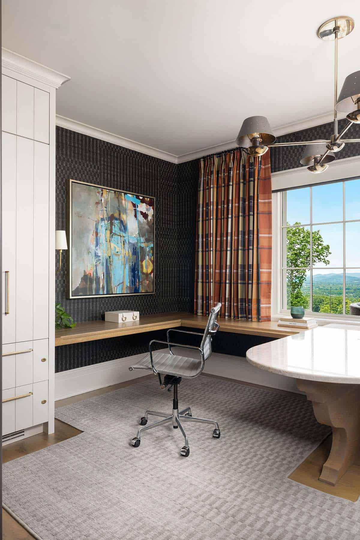 Home office with dark textured wallcovering, a floating wood corner desk, abstract painting, plaid drapes, grid window with mountain and treetop views, a chrome task chair, and a multi-arm pendant light fixture