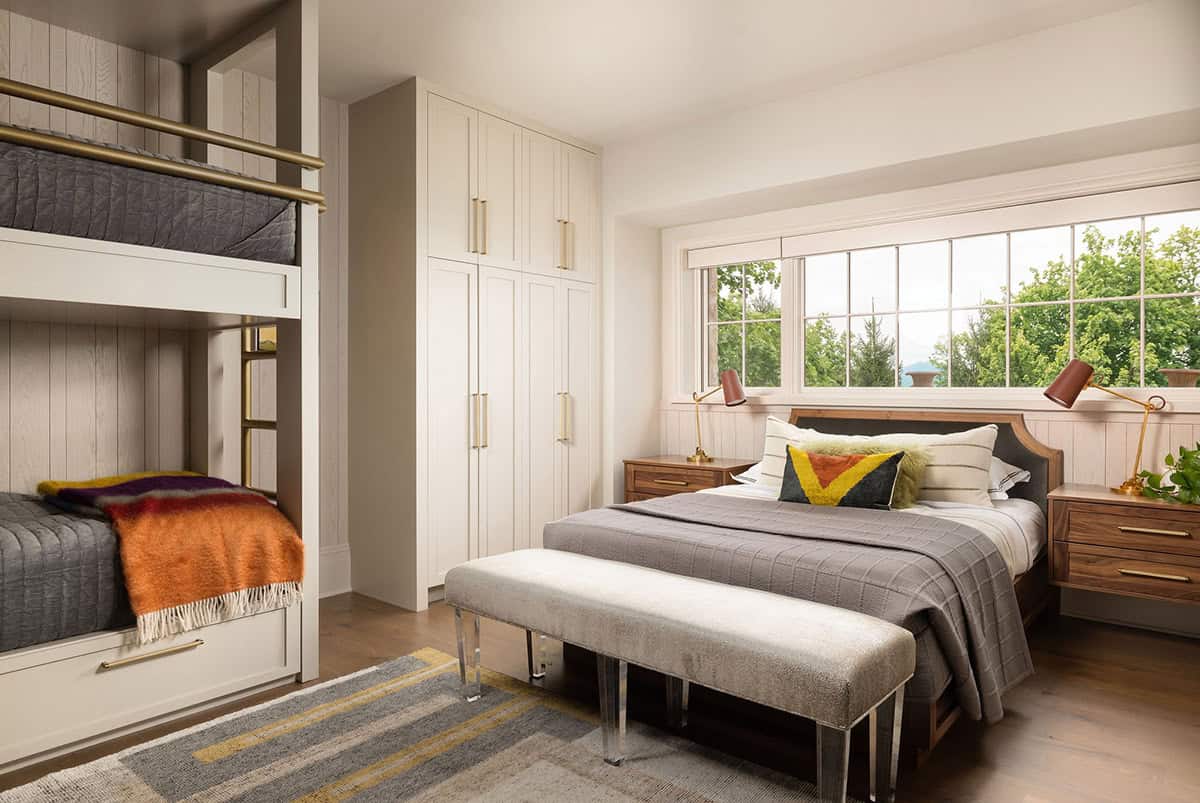 Guest bedroom with built-in bunk beds with brass safety rails and a trundle drawer, floor-to-ceiling white cabinetry with brass pulls, a walnut bed with upholstered headboard, walnut nightstands, and a grid window overlooking treetops