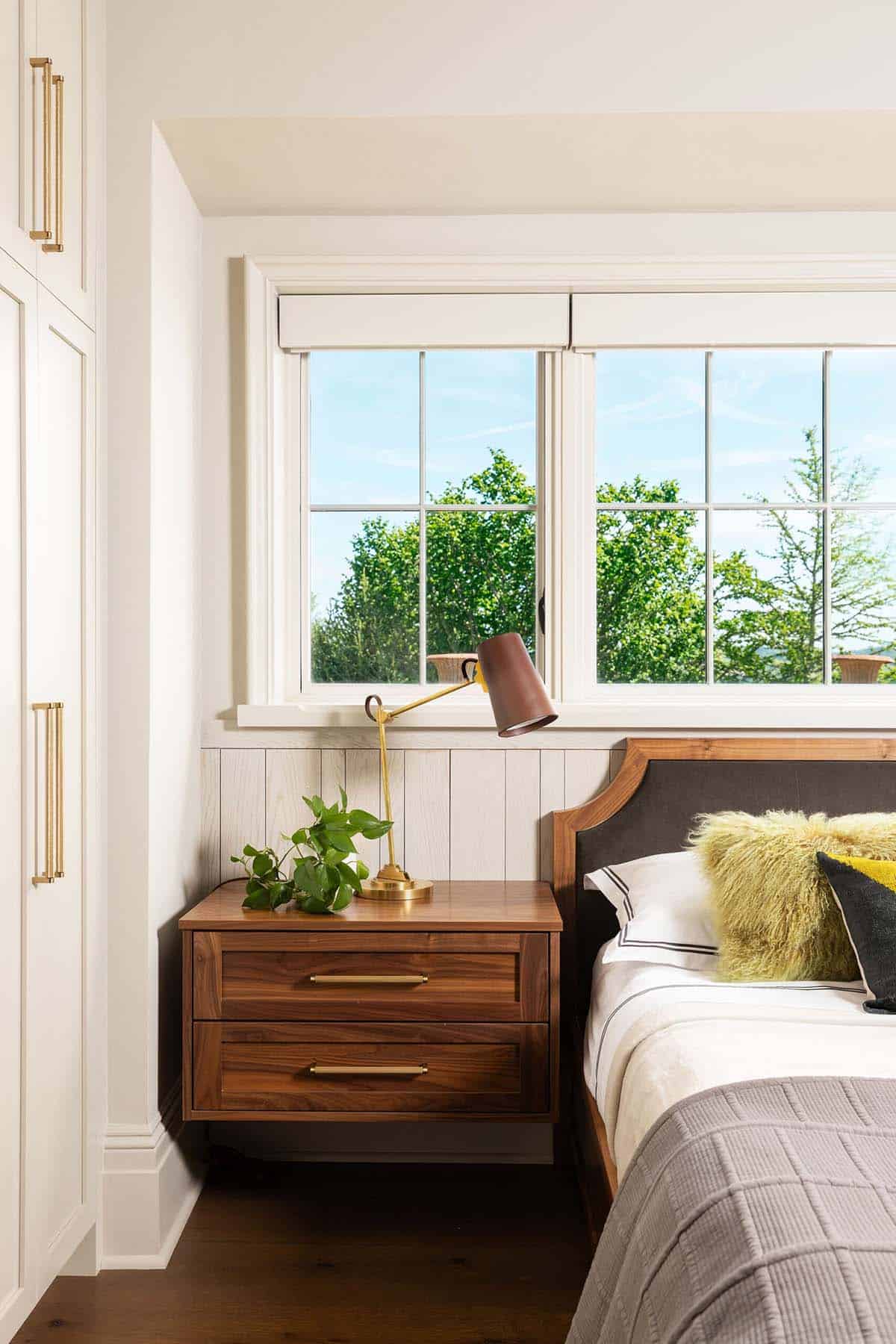 Close-up of a walnut two-drawer nightstand with brass pulls, a brass adjustable task lamp with a brown shade, trailing pothos plant, and a grid window overlooking green trees