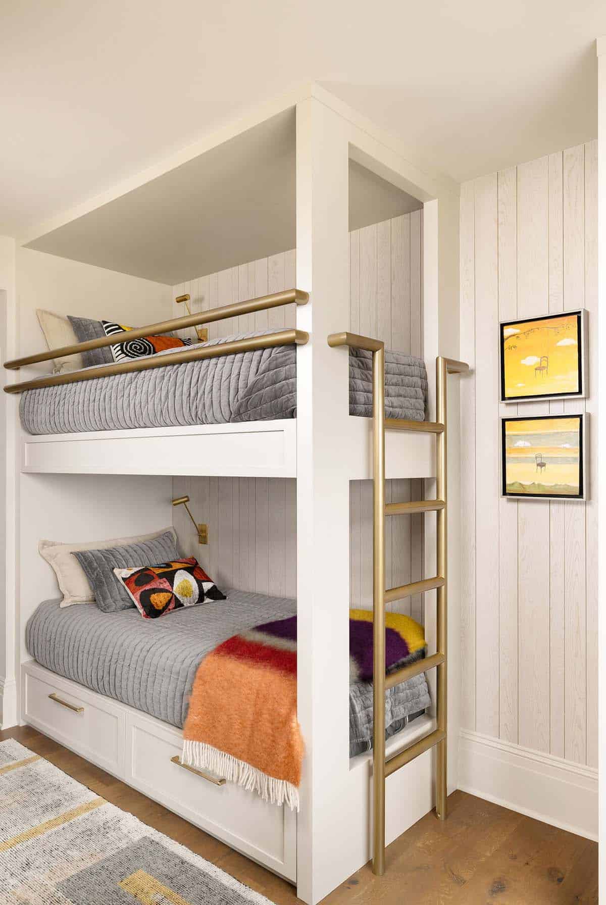 Built-in bunk beds with white painted frames, brass safety rails and ladder, shiplap-style paneled walls, colorful accent pillows and throw blankets, under-bed storage drawers, and two small framed yellow paintings