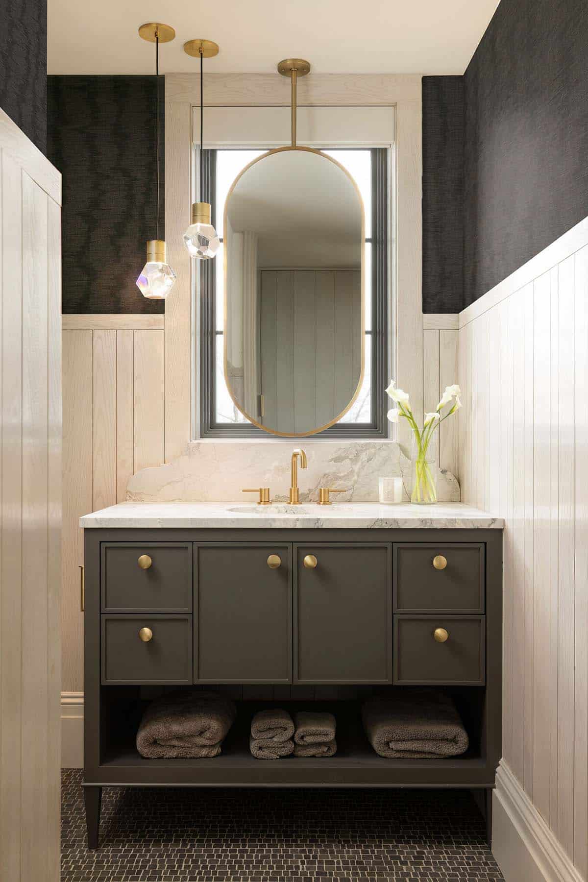 Bathroom vanity with a dark gray painted cabinet, brass knob hardware, marble countertop with a shaped backsplash, brass widespread faucet, oval brass-framed mirror, crystal geometric pendant lights, and dark mosaic tile floor