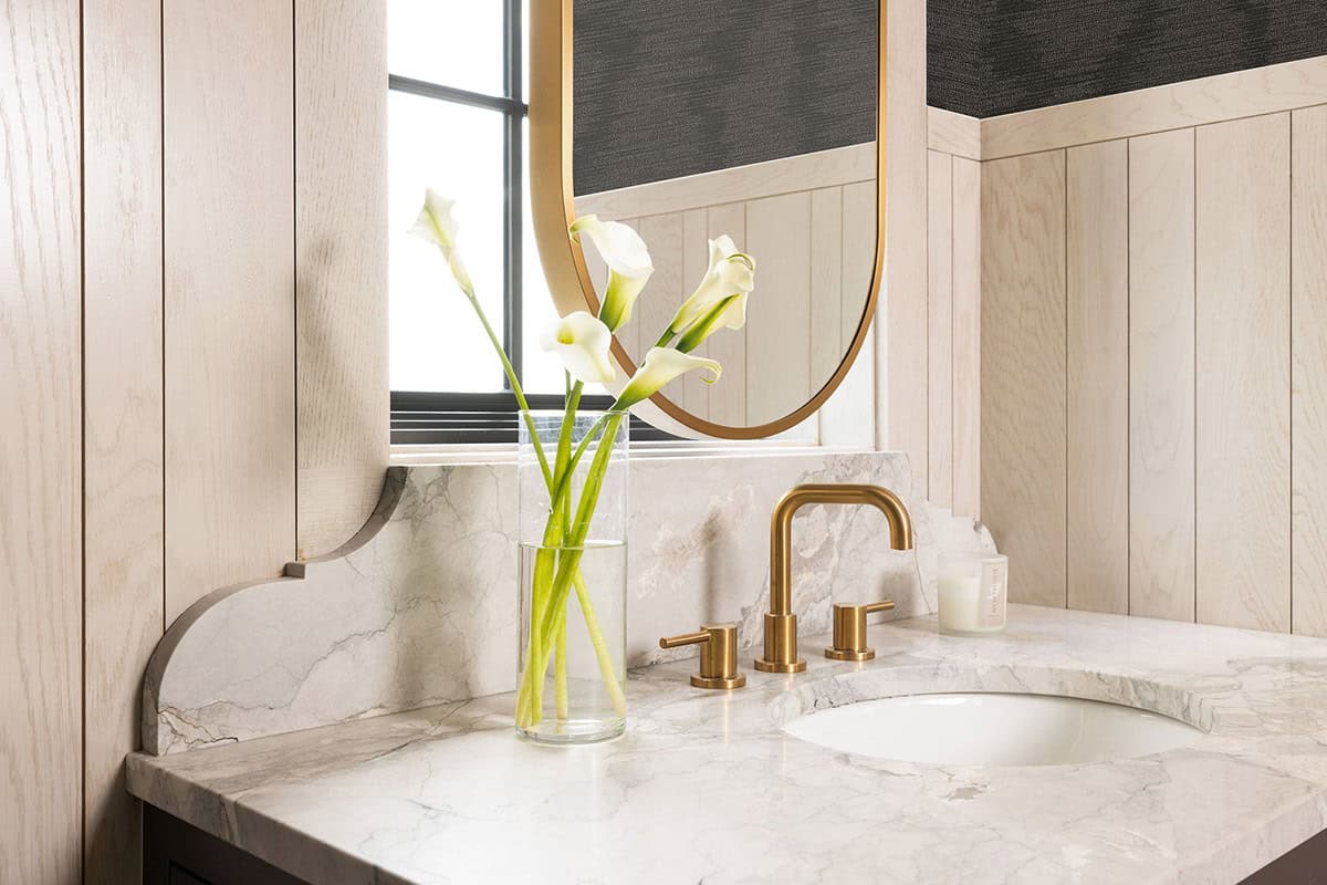 Close-up of a bathroom vanity with a marble countertop and shaped backsplash, brass widespread faucet, oval brass-framed mirror, and a clear glass vase of white calla lilies
