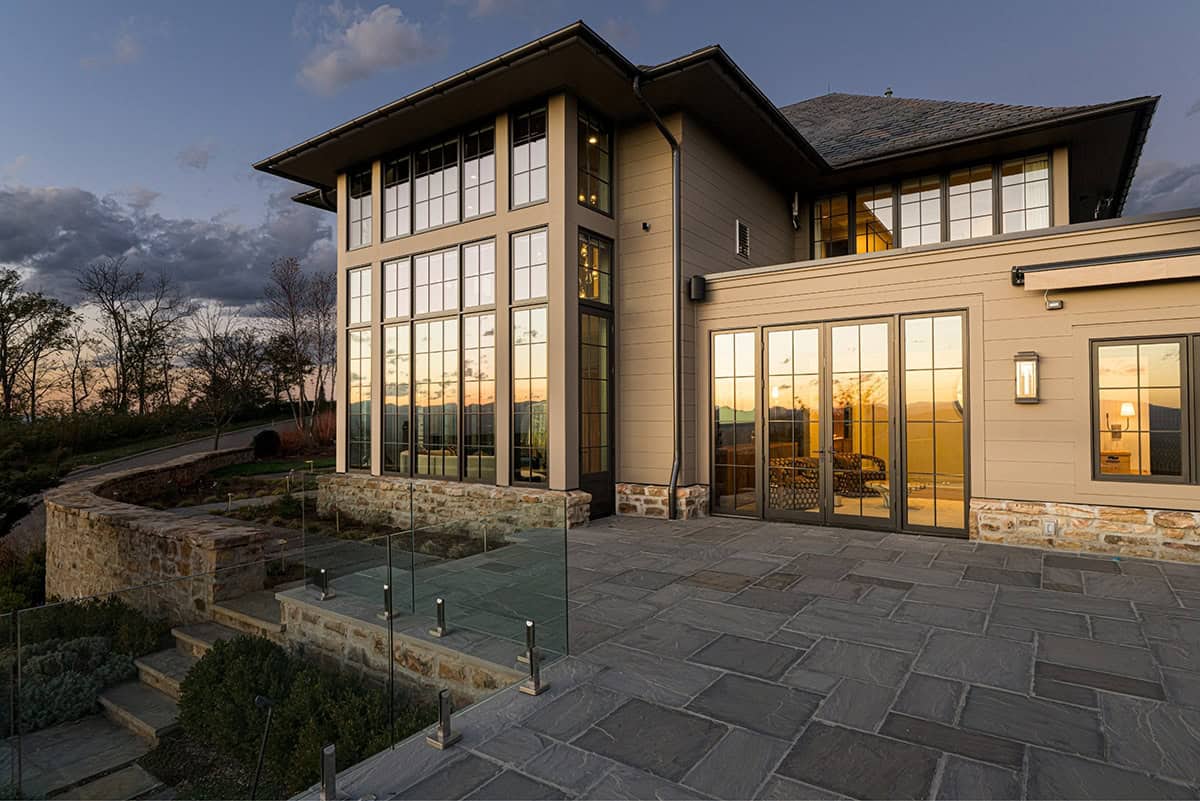 Rear exterior of the home at dusk showing two-story black-framed grid windows reflecting the sunset, a stone and lap siding facade, stone patio with glass panel railing, and layered terrace landscaping