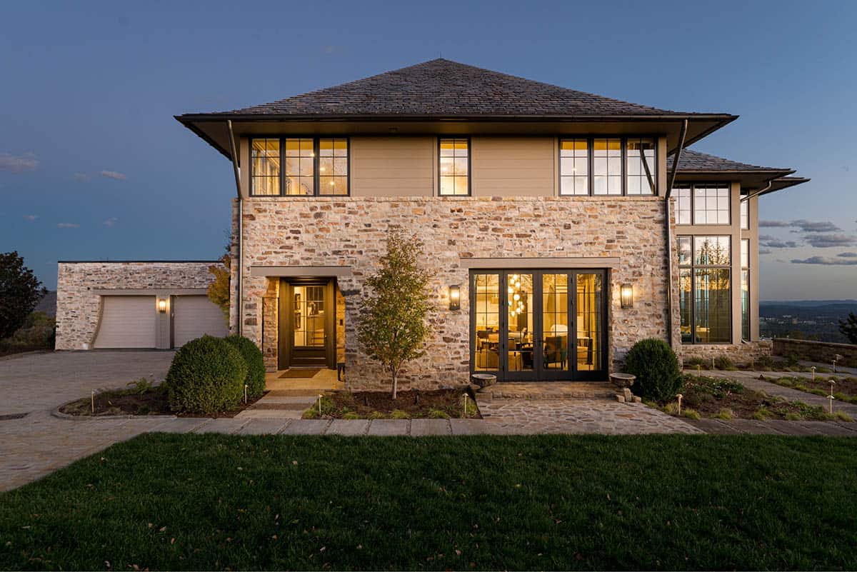 Front exterior at dusk with warm interior light glowing through large black-framed windows, a natural stone facade, flagstone entry walk, clipped boxwoods, and a single-car garage wing