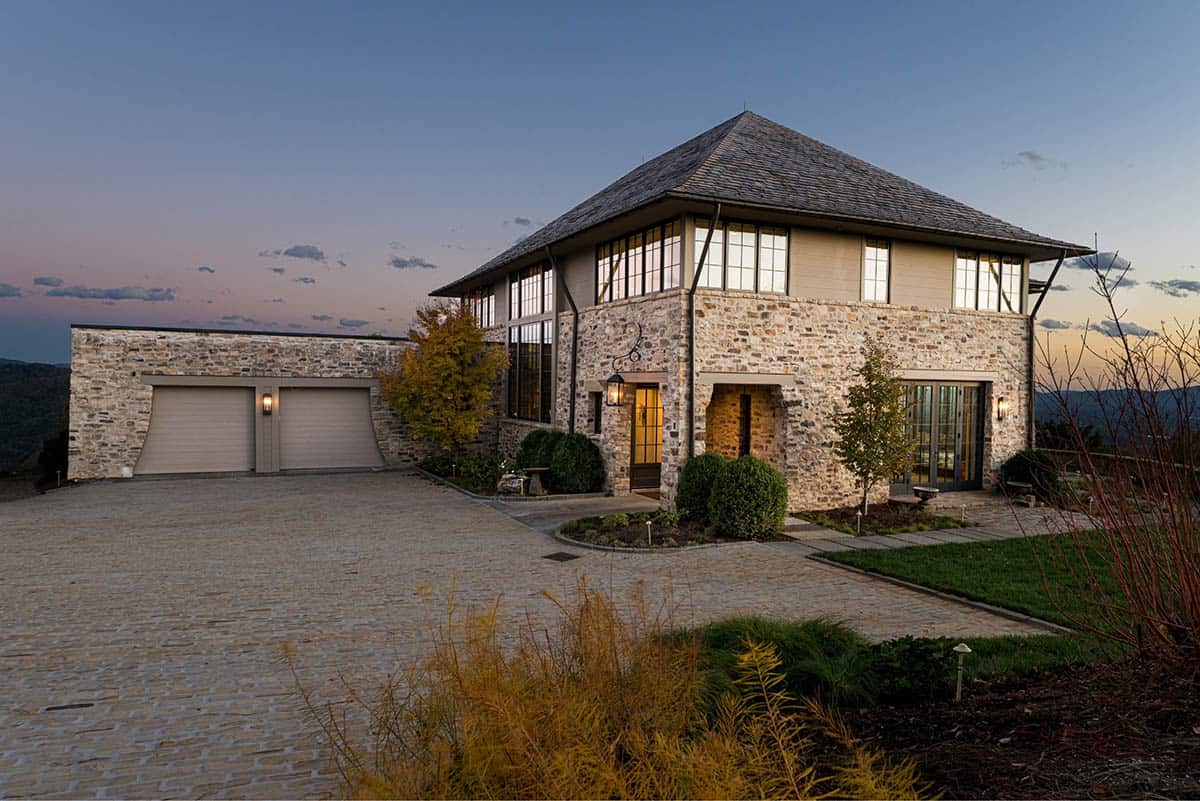 Dusk front exterior of the stone home with lit entry lanterns, two-car garage, cobblestone driveway, clipped boxwoods, tall grid windows, and a pink and blue sky with mountain silhouettes