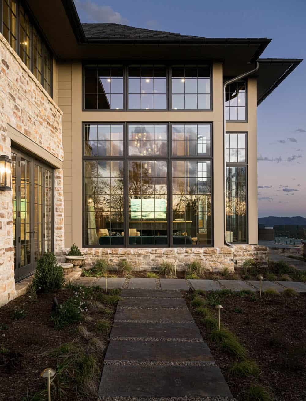 Nighttime exterior close-up of the double-height living room grid window wall, revealing the lit interior with the fireplace and chandelier, stone and siding facade, and a slate stepping stone path through mulched plantings