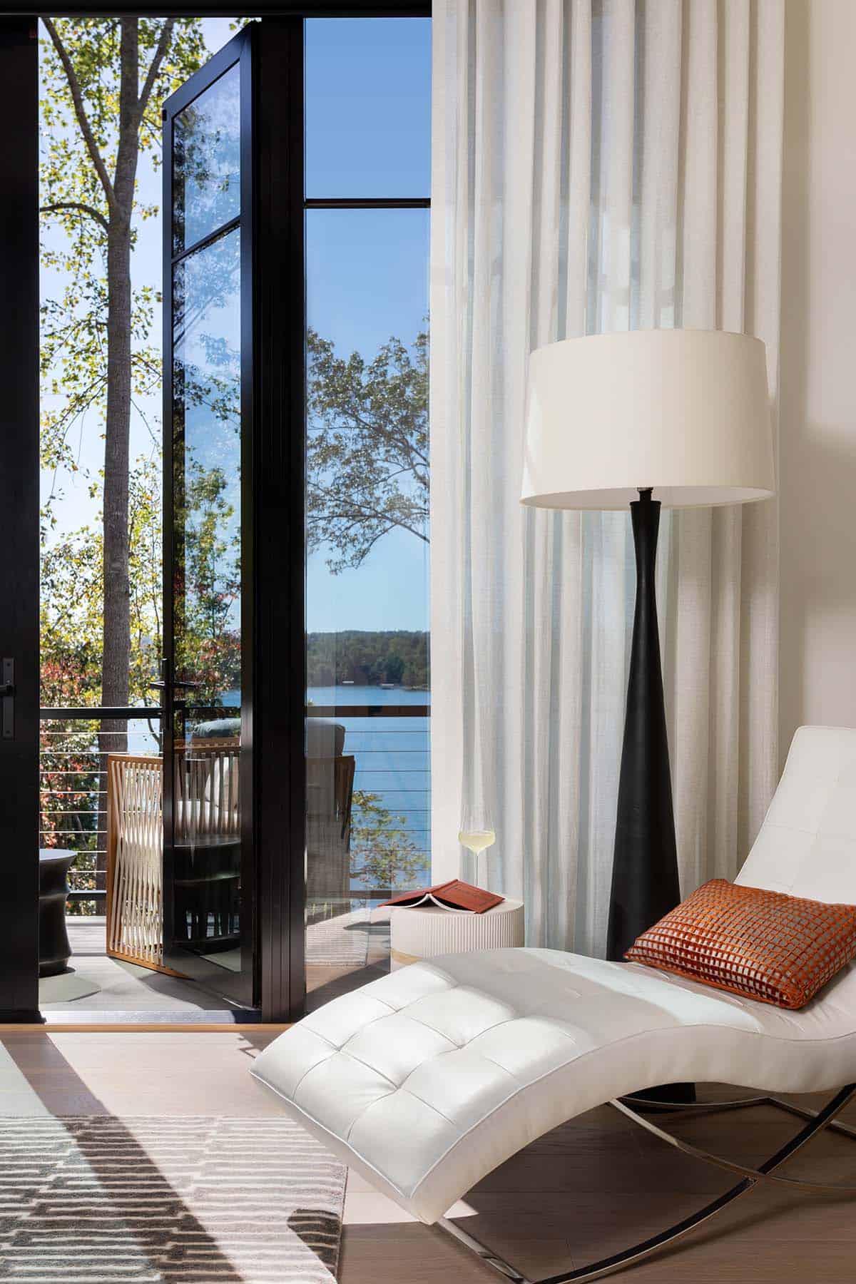 Primary bedroom with a white tufted chaise lounge beside floor-to-ceiling glass doors overlooking a lake