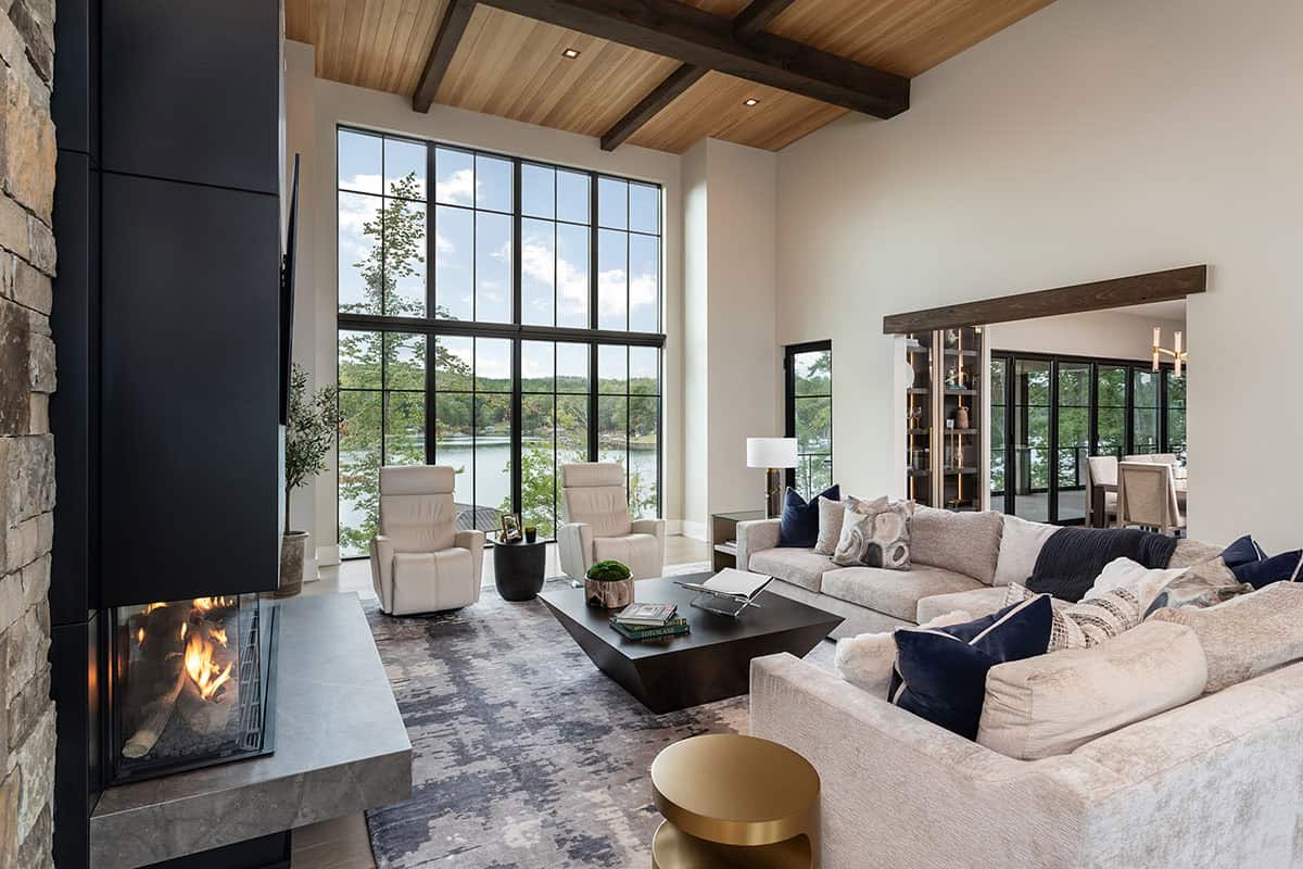 Vaulted great room with wood beam ceiling, two-sided stone fireplace, and floor-to-ceiling lake-view windows