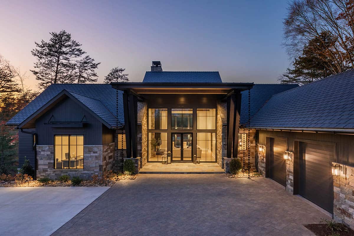 Front entry of lake house at dusk with illuminated two-story glass foyer, stone columns, and paver courtyard
