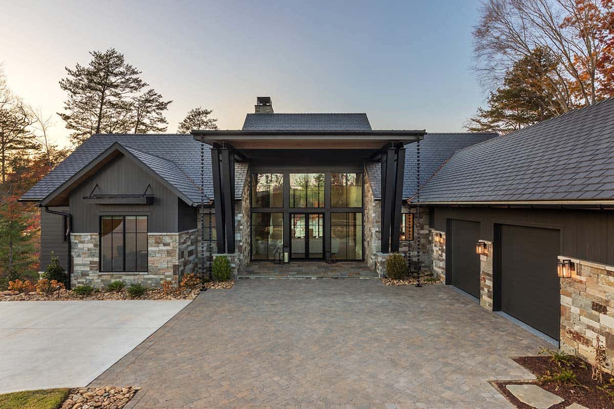 Daytime front facade of modern lake house with stone and dark siding, large glass entry, and paver driveway