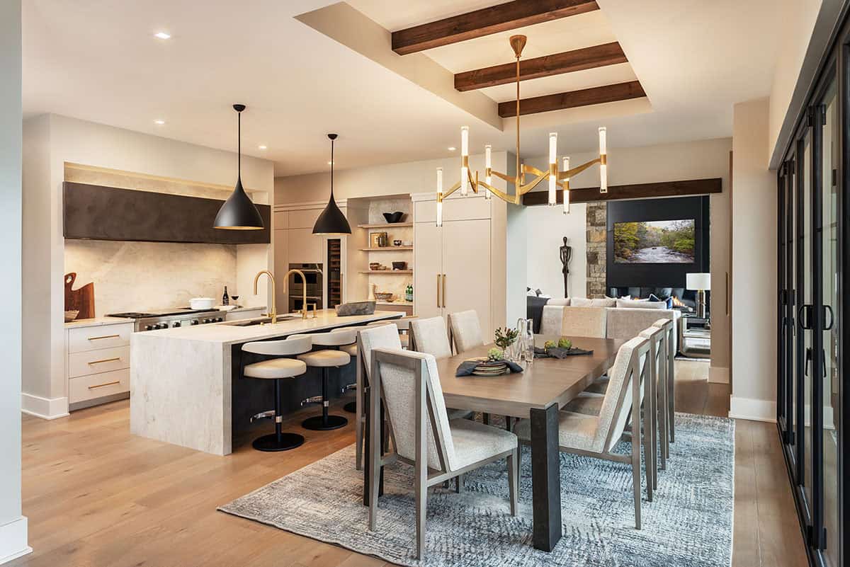 Open-plan dining and kitchen with brass chandelier, marble island, dark pendant lights, and wood beam ceiling