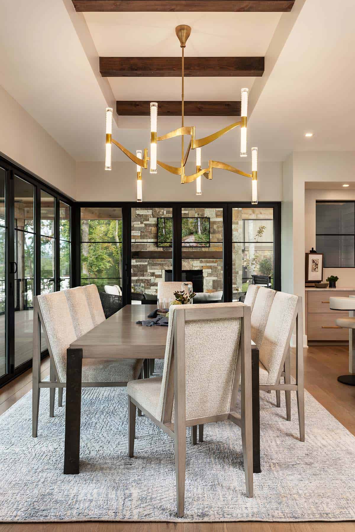 Dining room with sculptural brass chandelier, gray upholstered chairs, and view to outdoor stone fireplace
