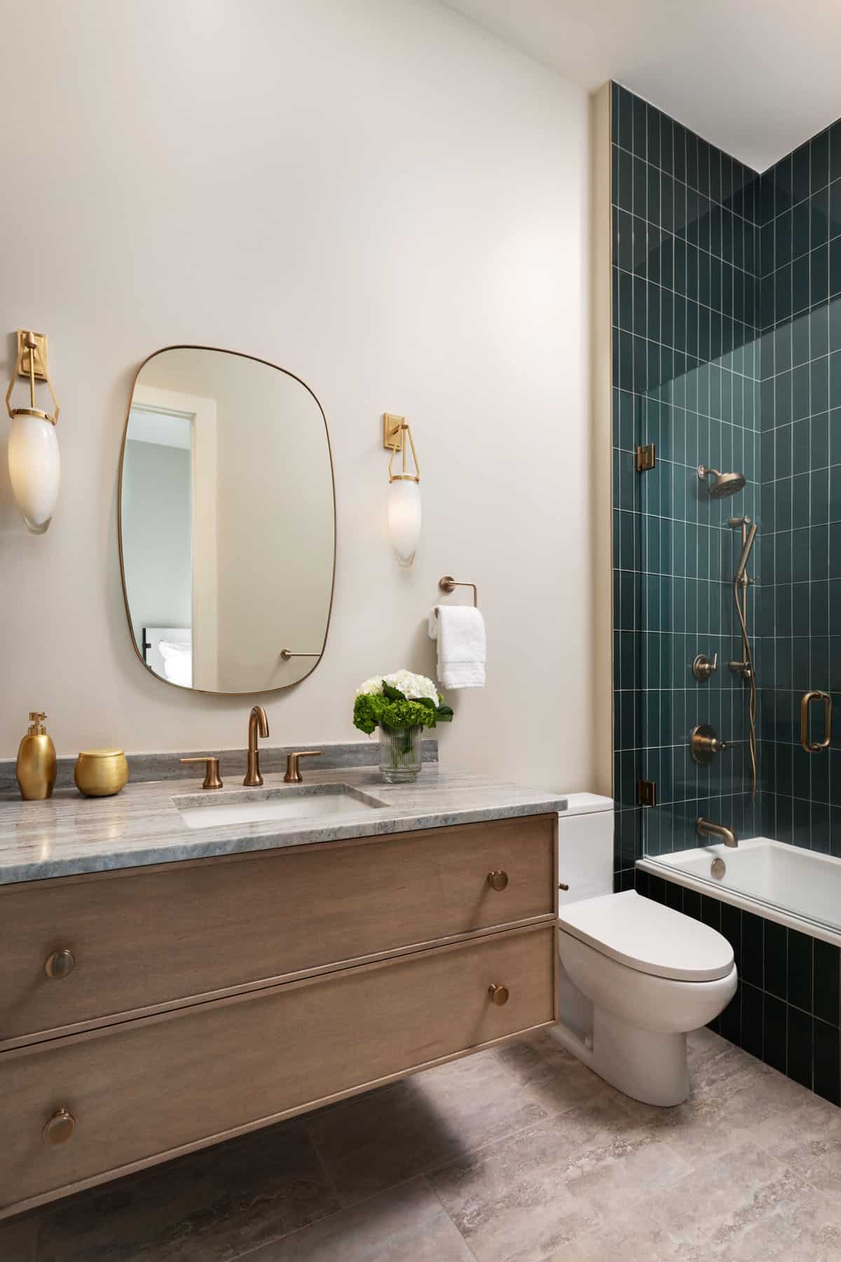 Guest bathroom with deep teal vertical tile shower, brass fixtures, floating wood vanity, and oval mirror