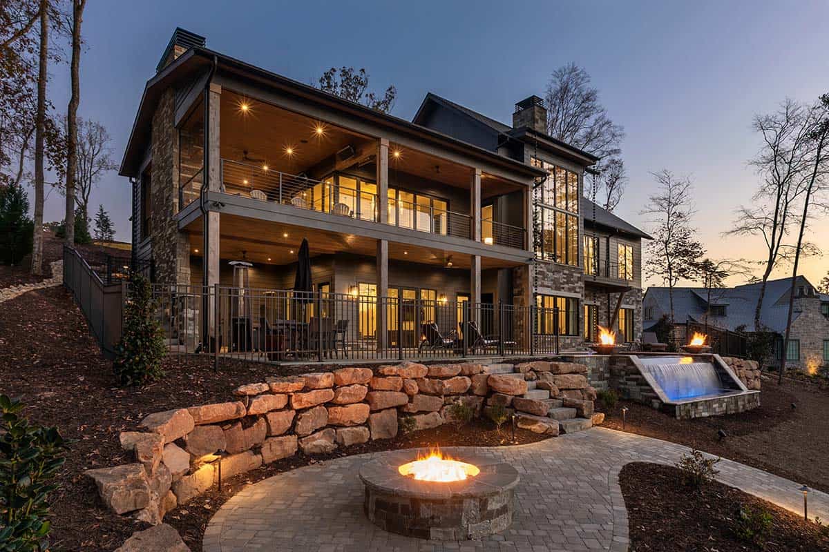 Lake house rear exterior at dusk with multi-level decks, fire pit, water feature, and stone retaining walls