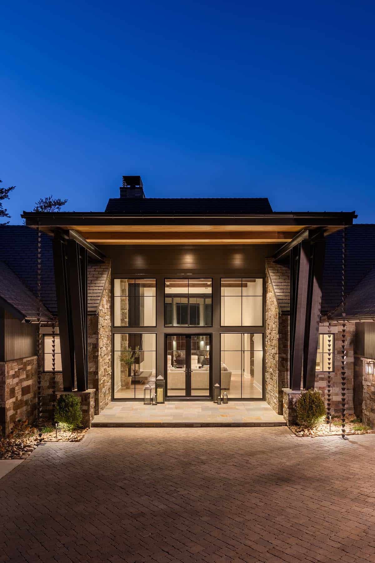 Lake house entry portico at night with two-story glass facade, stone columns, wood soffit, and paver forecourt