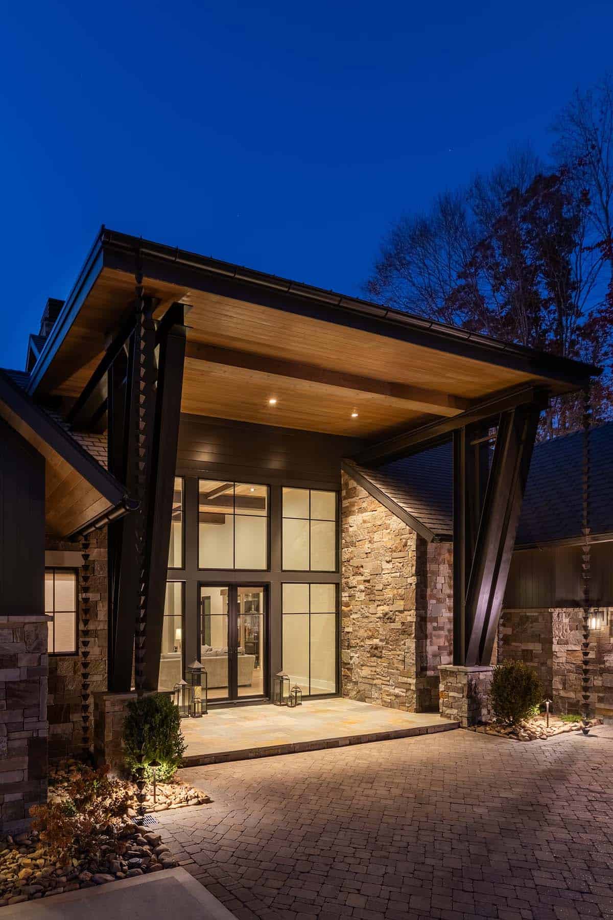 Angled night view of lake house entry with dark steel beams, warm wood ceiling, and illuminated glass foyer