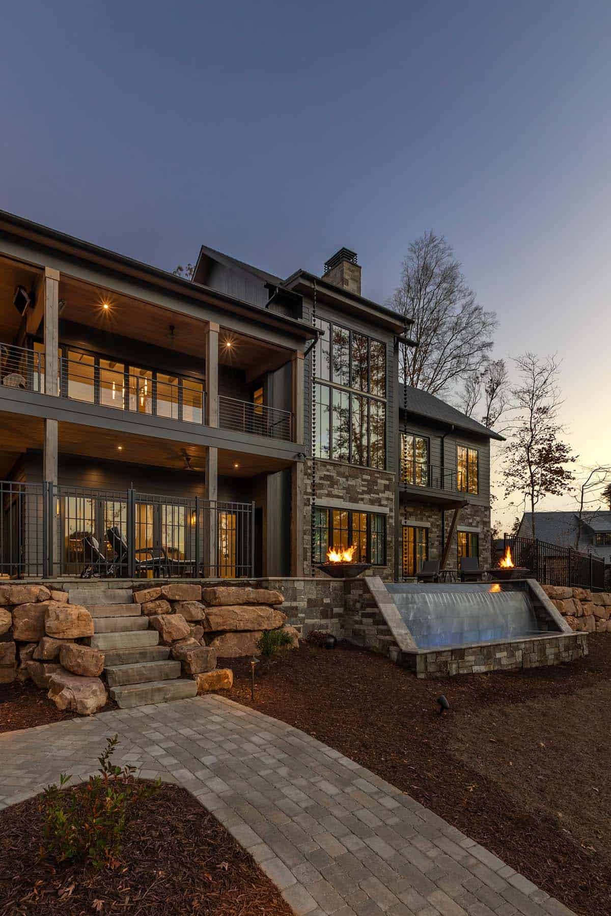 Lake house rear at dusk showing stacked covered porches, cascading waterfall feature, and fire bowl flanking stone wall