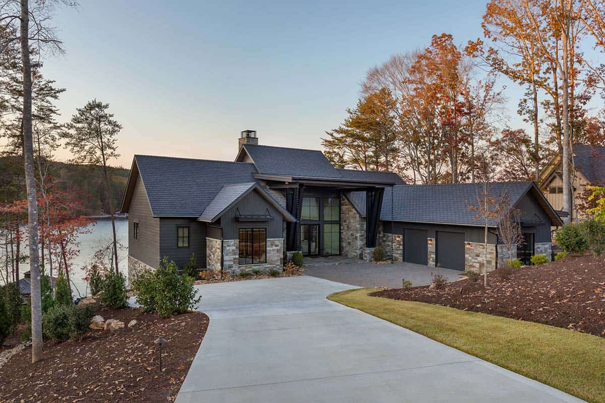 Wide daytime view of lake house front with dark siding, stone accents, two-car garage, and lake visible to the left