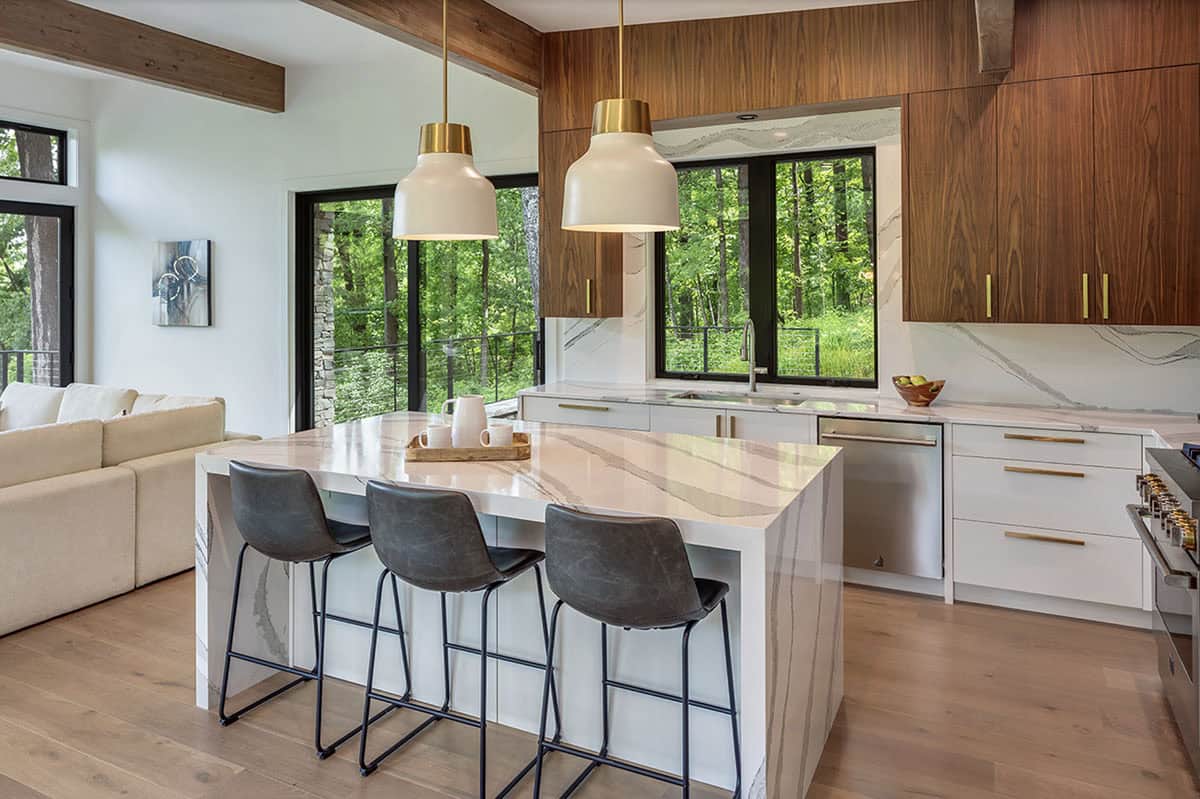 Modern kitchen with marble island, walnut cabinetry, brass pendant lights, and forest views