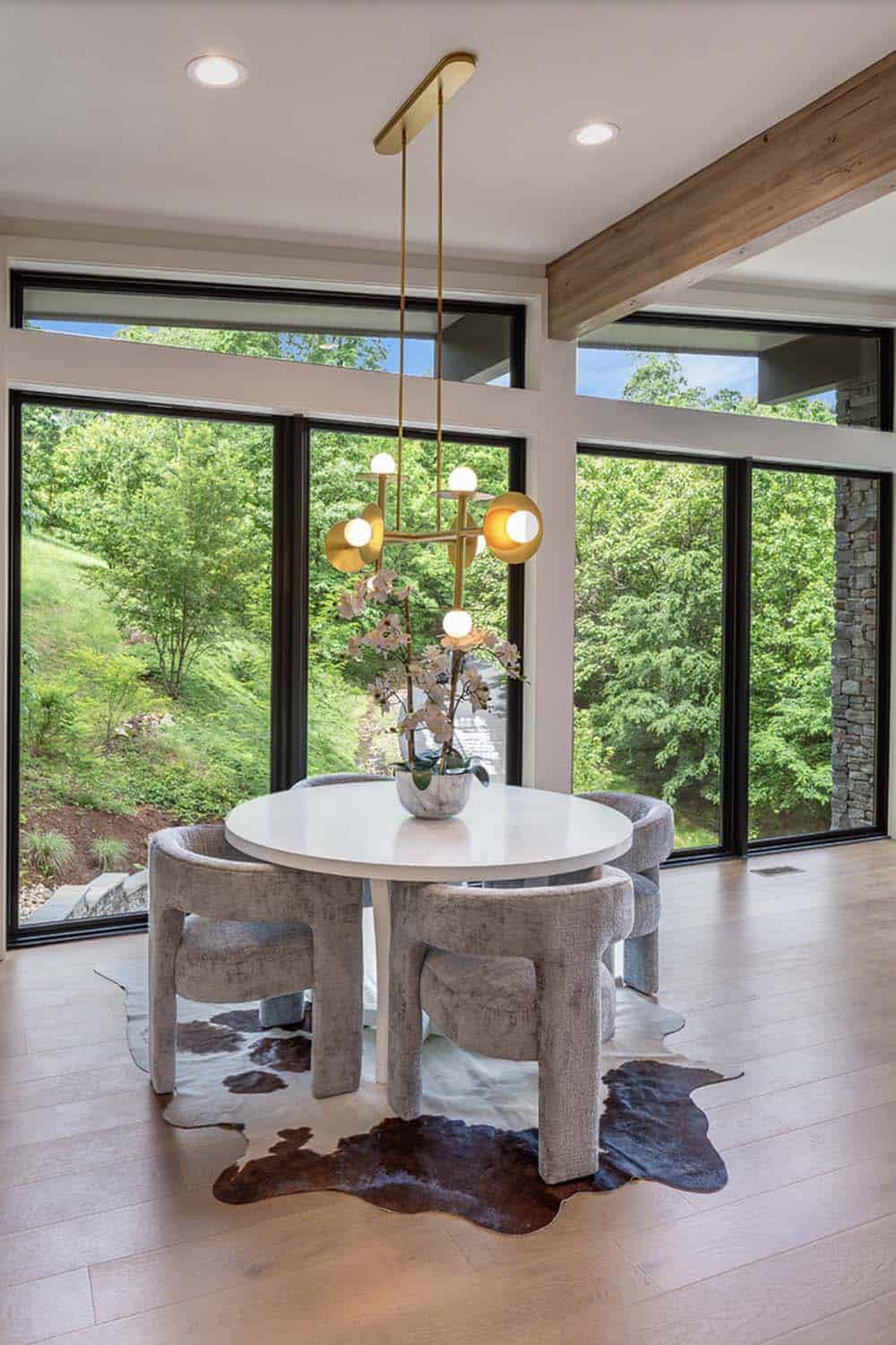 Round dining table with upholstered chairs beneath a brass sculptural chandelier, floor-to-ceiling windows behind