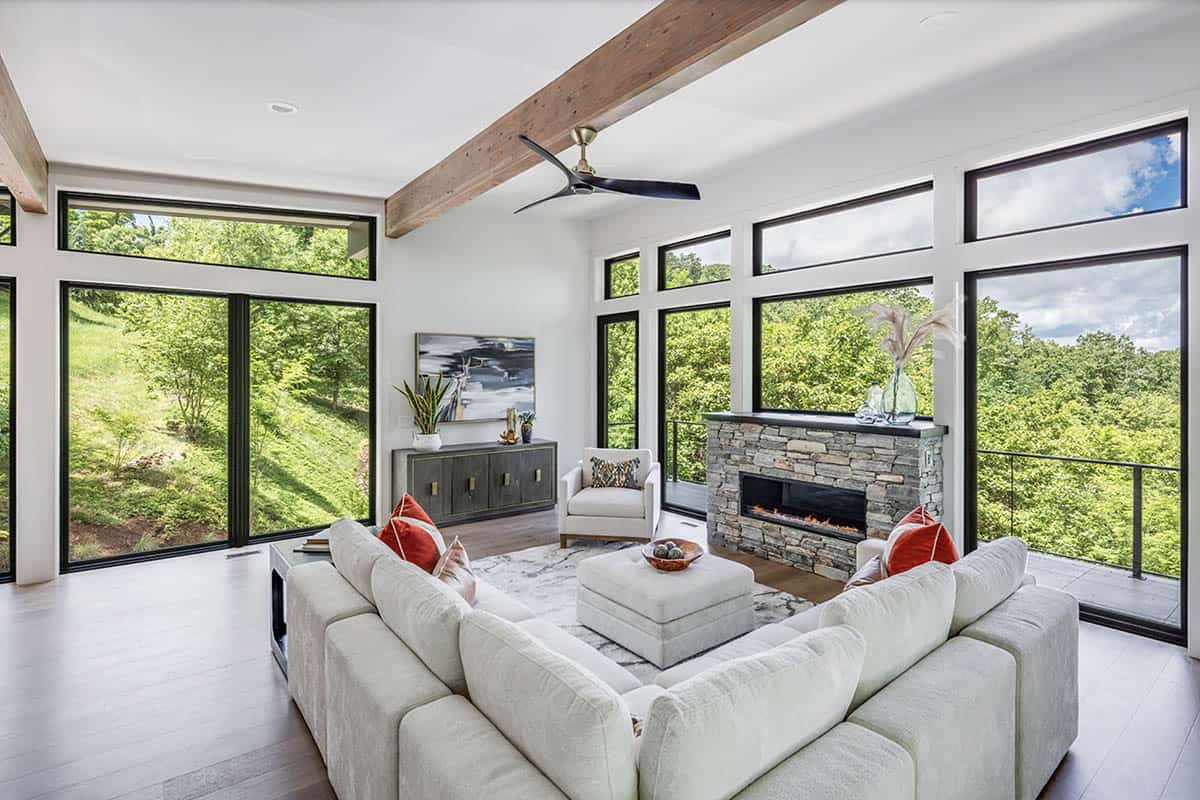 Living room with white sectional sofa, stone fireplace, and tall windows overlooking a forested hillside