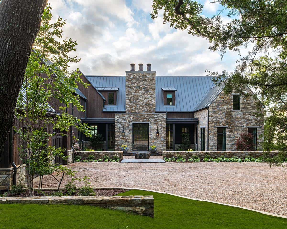 Stone and dark wood lake house with steep gabled metal roof, central stone chimney, and gravel motor court framed by a lush lawn