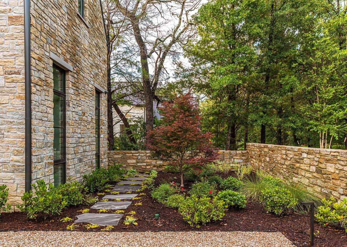 Side garden path of stone pavers leading along a stone exterior wall, lined with shrubs, a red Japanese maple, and a low stone garden wall