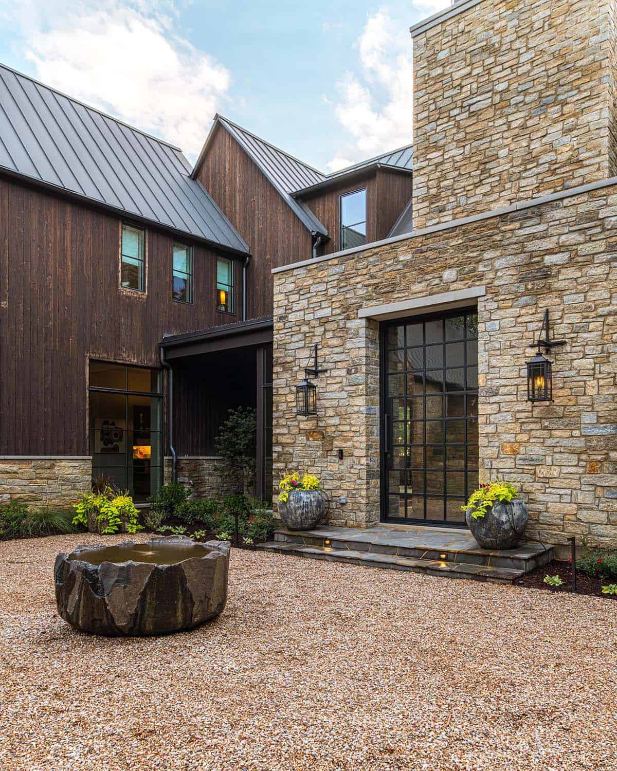 Stone entry tower with glass door and lantern sconces beside a carved stone water basin on a gravel courtyard